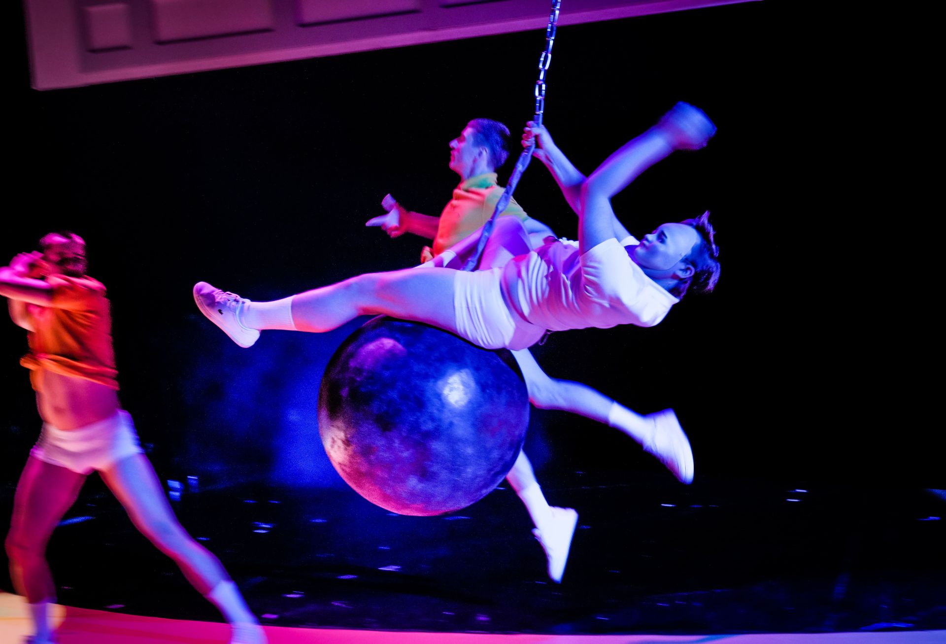 Circus performers on a large spherical prop under blue-pink stage lights; one person swings on a chain while others move dynamically in white and neon outfits.