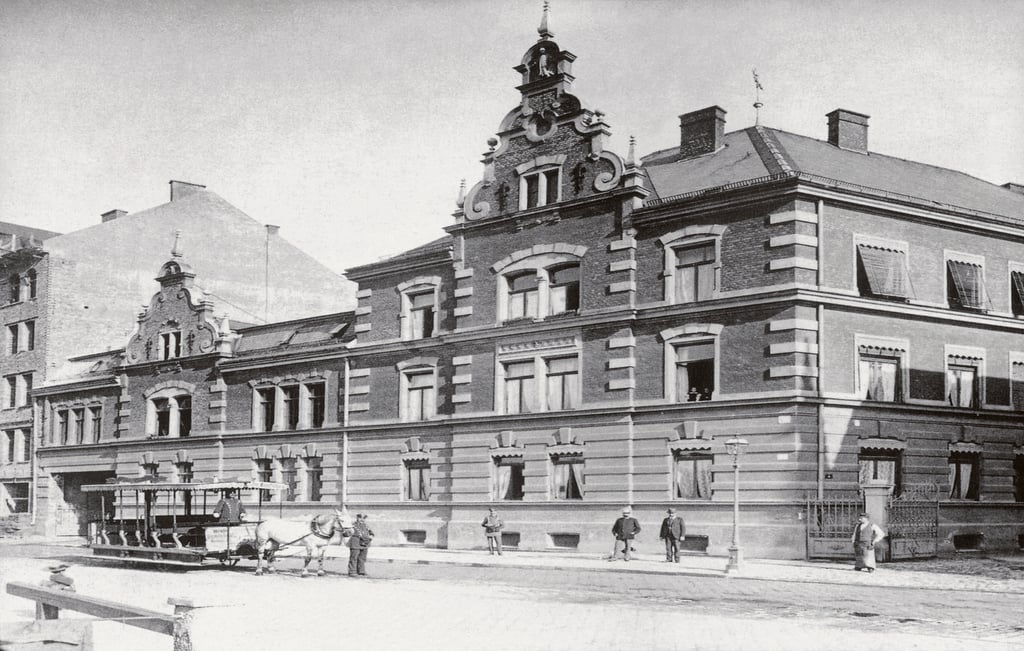 Historic brick building with ornate gable facade on a city street; horse-drawn cart in front and pedestrians nearby.