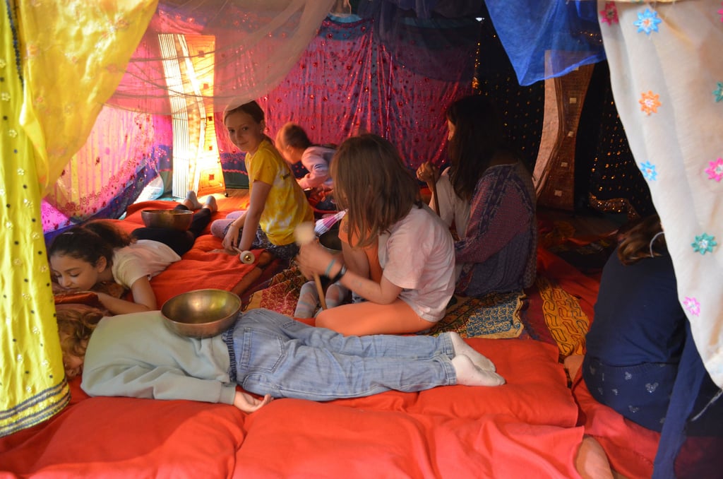 Children lie on red cushions inside a bright fabric canopy, playing with bowls and small percussion instruments as others watch.