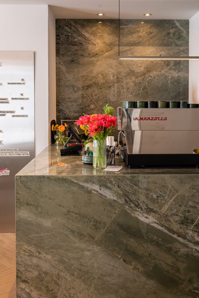 Cafe counter with a metallic La Marzocco espresso machine, pink flowers in a vase, and a marble backdrop.