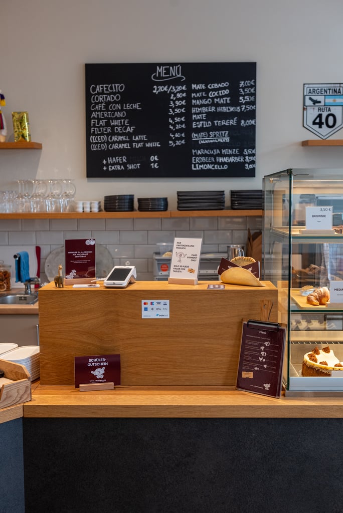 Cafe counter inside a cafe with a large chalkboard menu mounted on the wall behind, wooden shelves, and glass pastry display on the right.