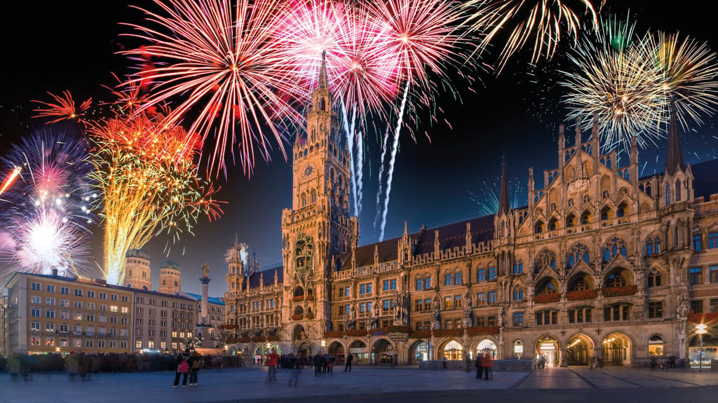 Fireworks explode over a historic Gothic town hall in a city square at night, with crowds watching below.