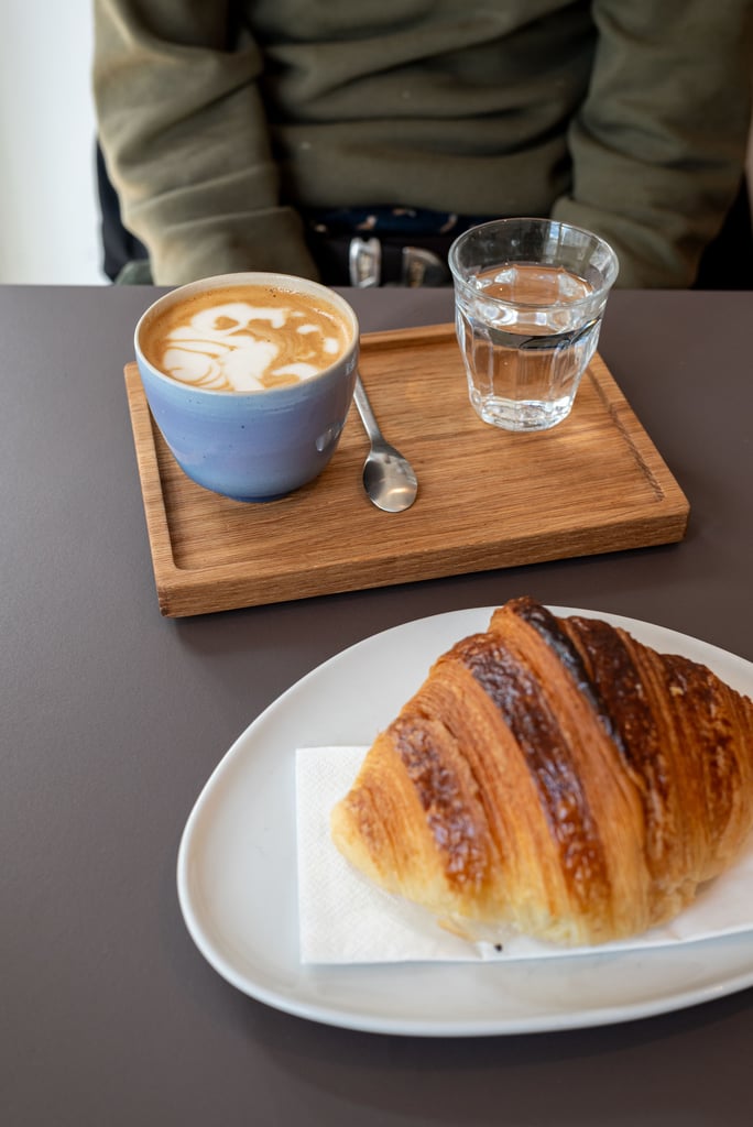 Blue ceramic mug with latte art on a wooden tray, next to a glass of water and a croissant on a plate in the foreground.