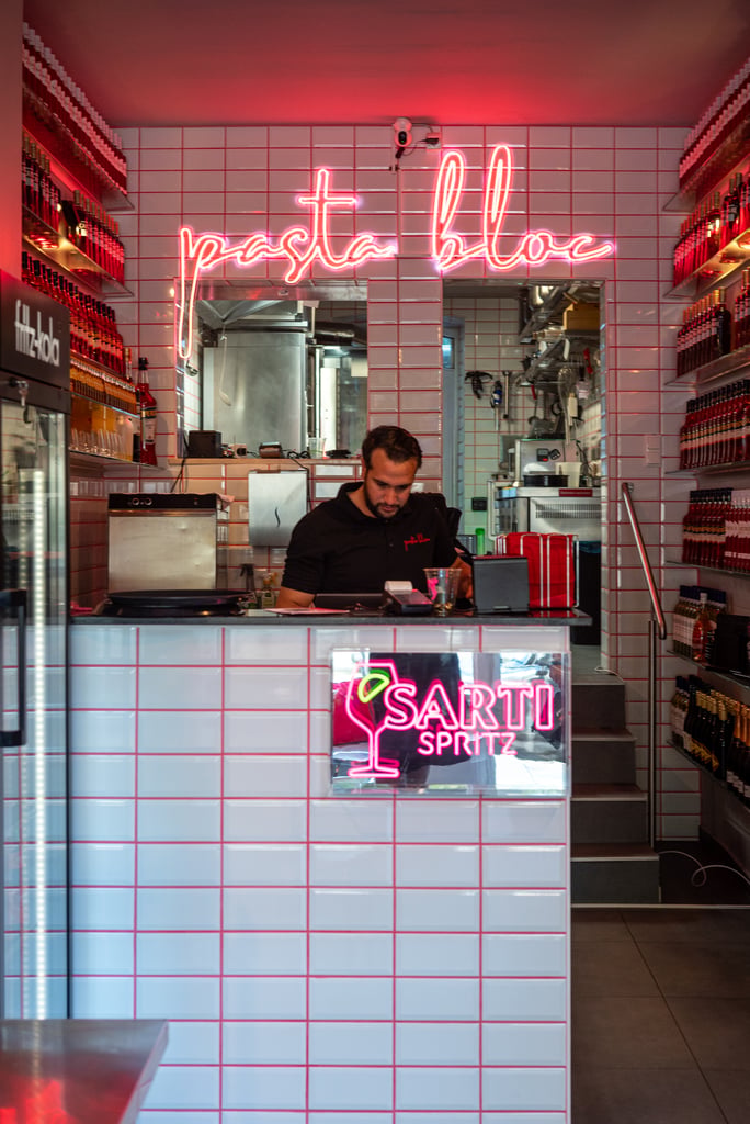 Interior of a pasta bar with white tile walls and a pink neon sign reading 'pasta' above the open kitchen, with a staff member behind the counter.