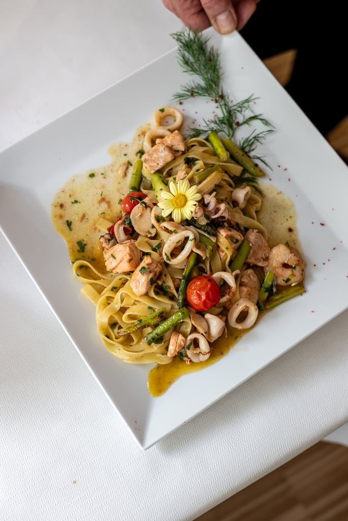 Creamy pasta with salmon, asparagus, and cherry tomatoes on a square plate, garnished with dill and a yellow flower.
