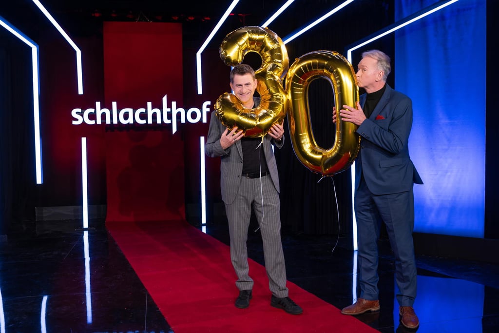Two men in suits hold large gold balloons '3' and '0' on a red carpet, neon-lit stage with 'schlachthof' sign in the background.