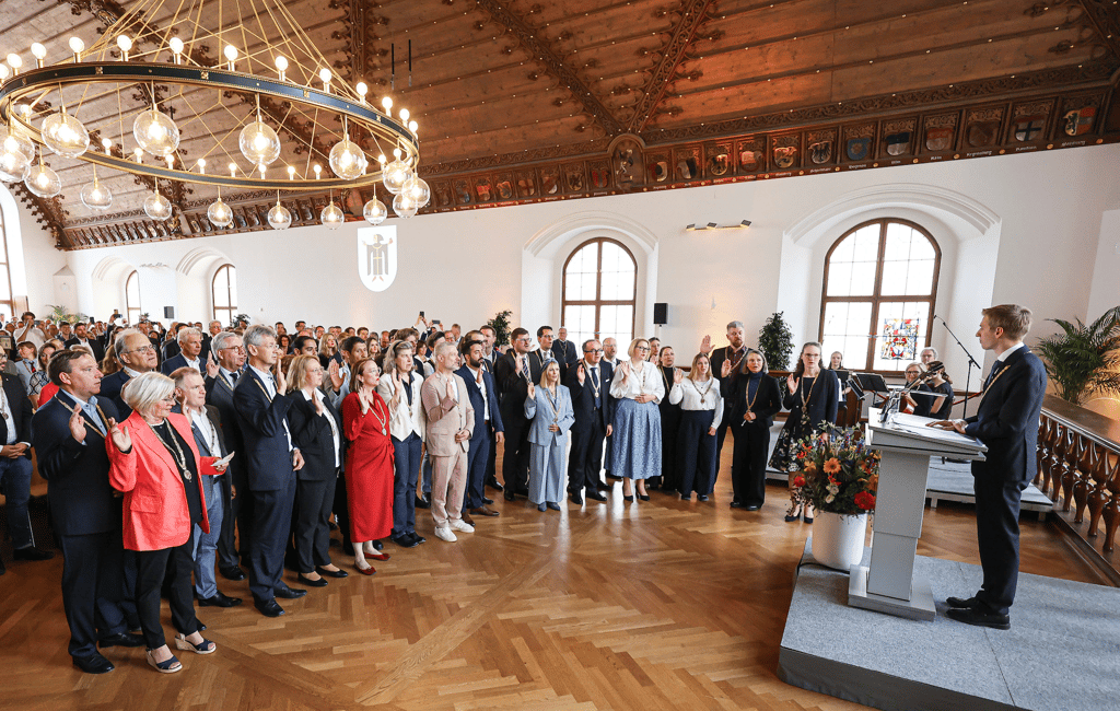 Formal ceremony in a wood-paneled hall; a speaker stands at a lectern as a large audience raises their hands in oath or pledge, with a grand chandelier overhead.