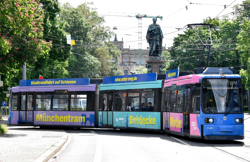 Colorful Munich tram with blue and purple panels on a sunny street, statue in the background and overhead wires above.