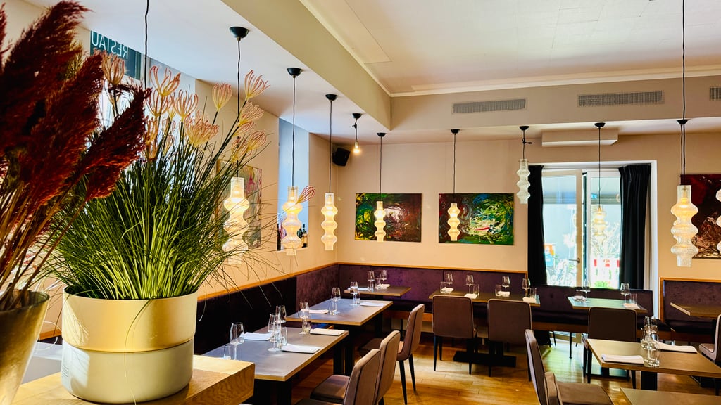 Restaurant dining area with tall potted plants in the foreground, tables set with wine glasses and napkins.