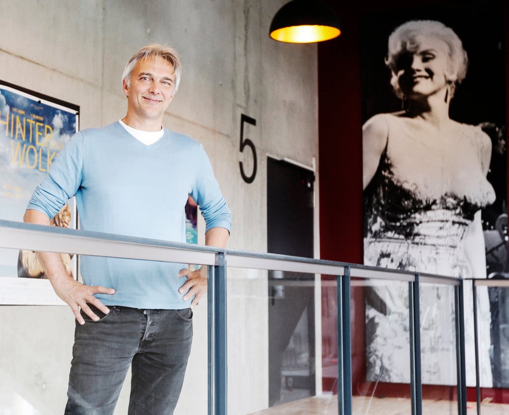 Smiling man in a light blue long-sleeve shirt stands with hands on hips beside a glass railing indoors, with a large black-and-white portrait on the wall behind him.