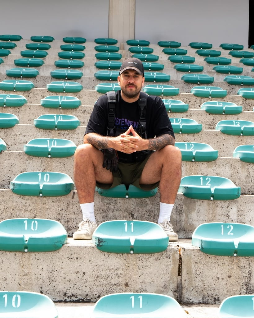 Man seated on stadium bleachers, wearing a black shirt and backpack, hands clasped, facing camera.