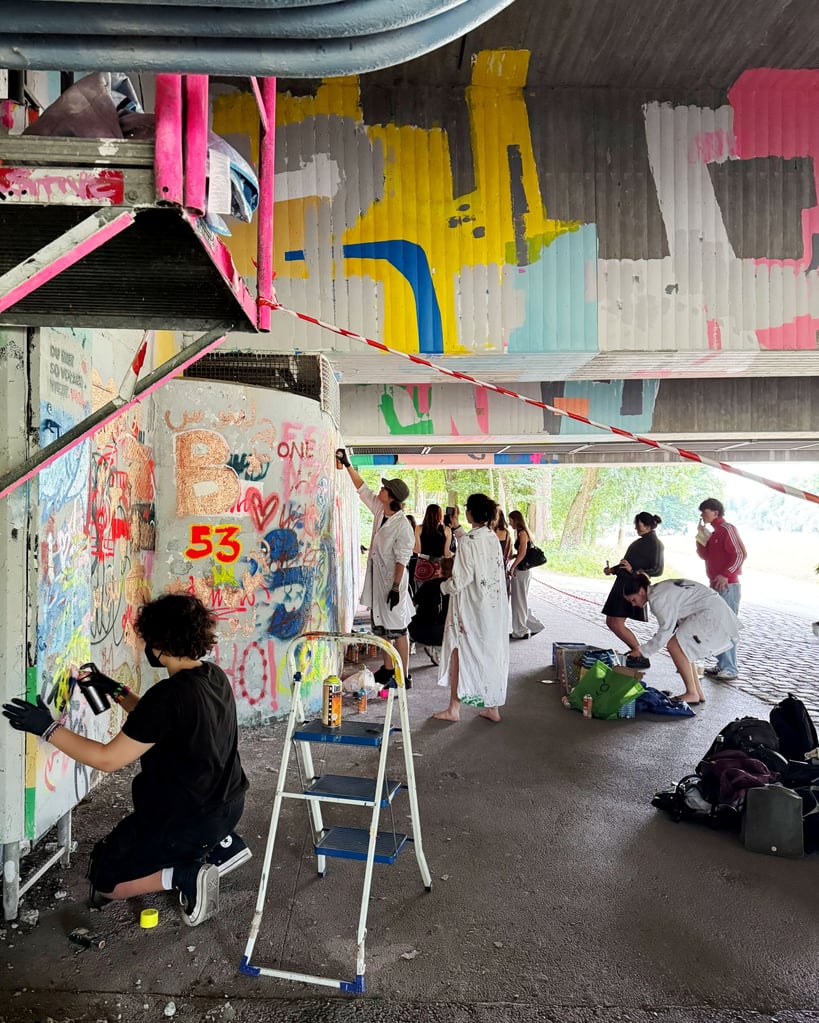 Group of people painting a colorful graffiti mural under a concrete overpass, with ladders, paint cans, and bags on the ground.