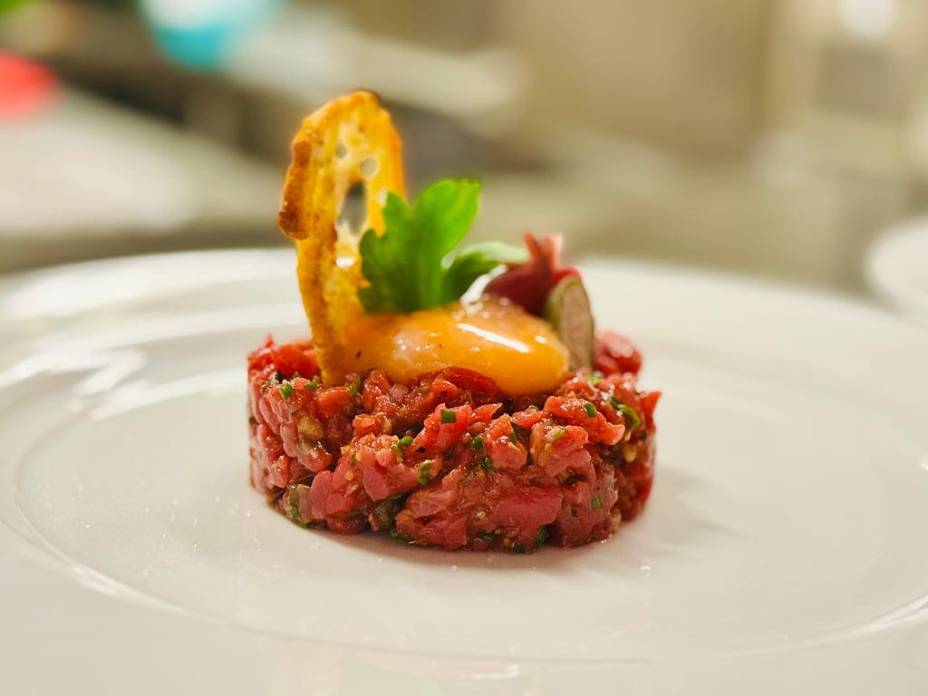Beef tartare formed into a small tower, topped with a crispy chip and a parsley leaf, around a red sauce on a white plate.