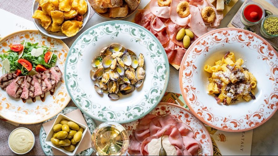 A colorful Italian antipasto spread with clams, roasted potatoes, olives, sliced meat, salad, and pasta on decorative plates.