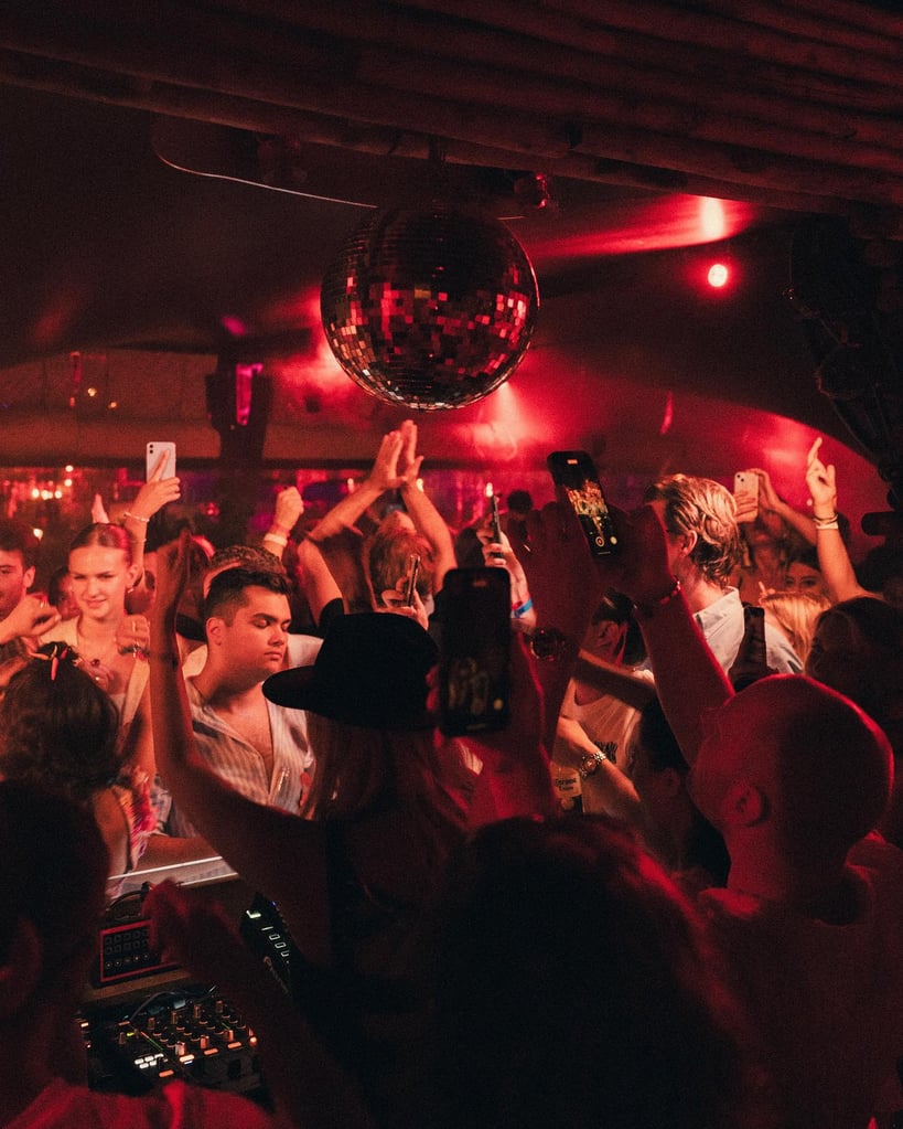 Crowded nightclub dance floor under red lights with a mirrored disco ball overhead and people raising smartphones to record the moment.