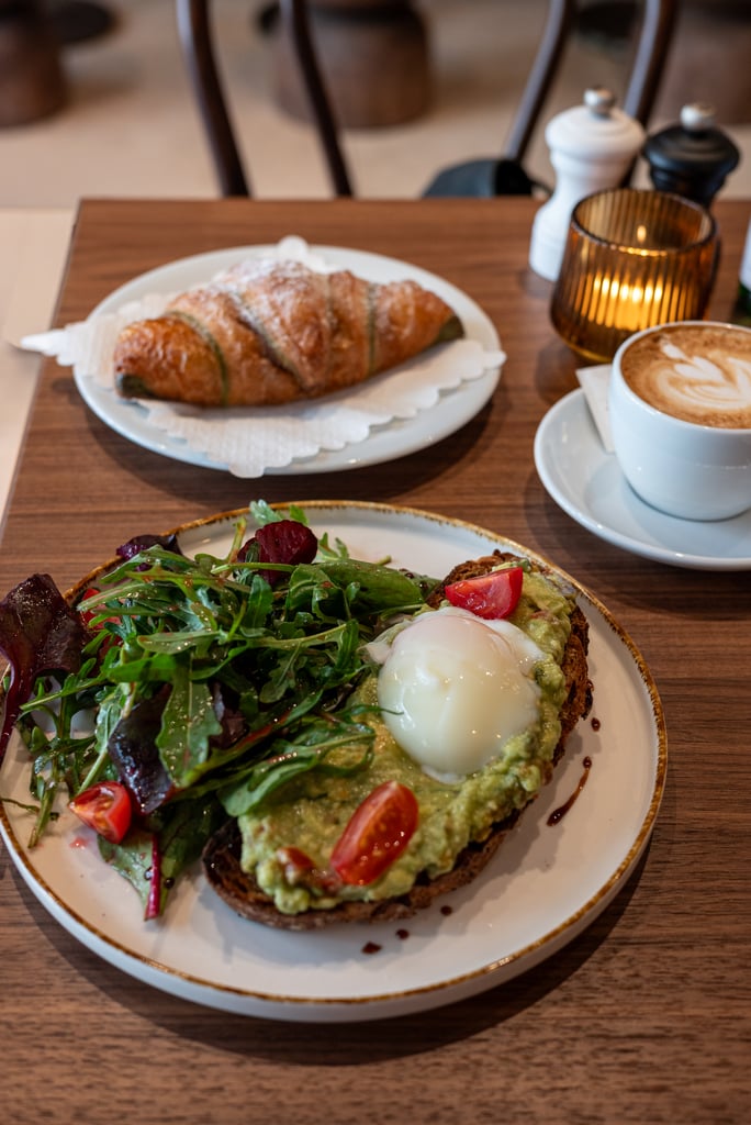 Avocado toast with greens and cherry tomatoes and a soft-cooked egg on a plate; coffee cup and a croissant in the background on a wooden table.