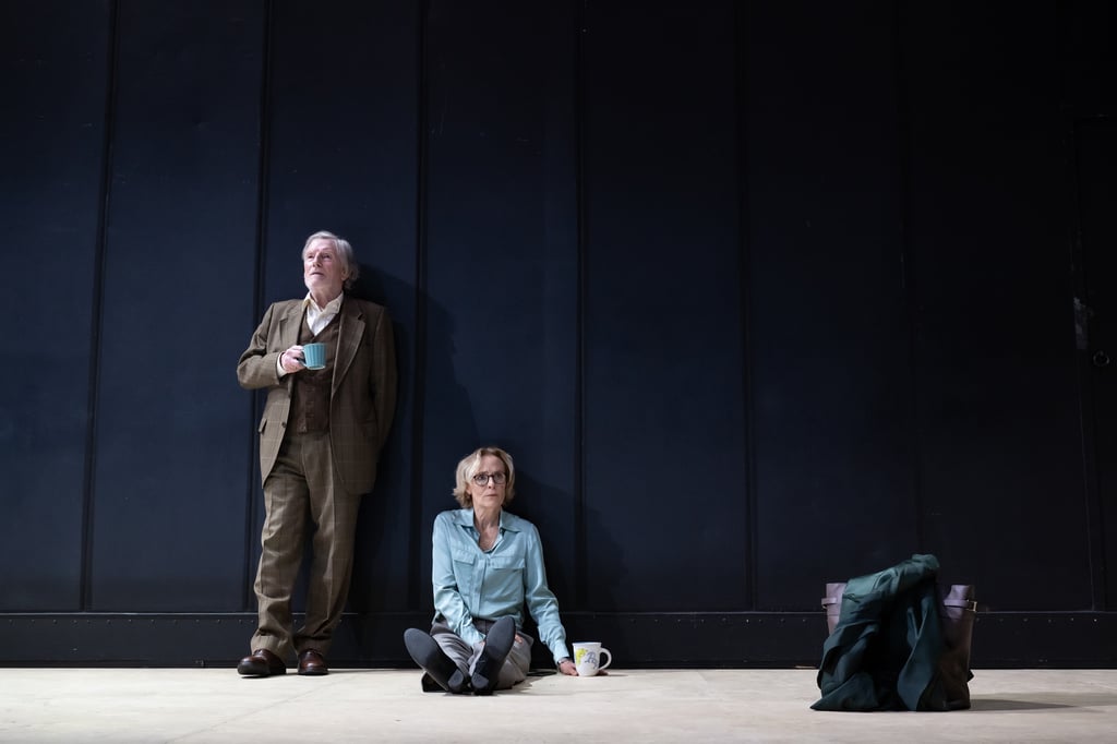An older man in a brown plaid suit leans against a dark wall, holding a turquoise mug, with a woman in a light blue shirt seated on the floor nearby.