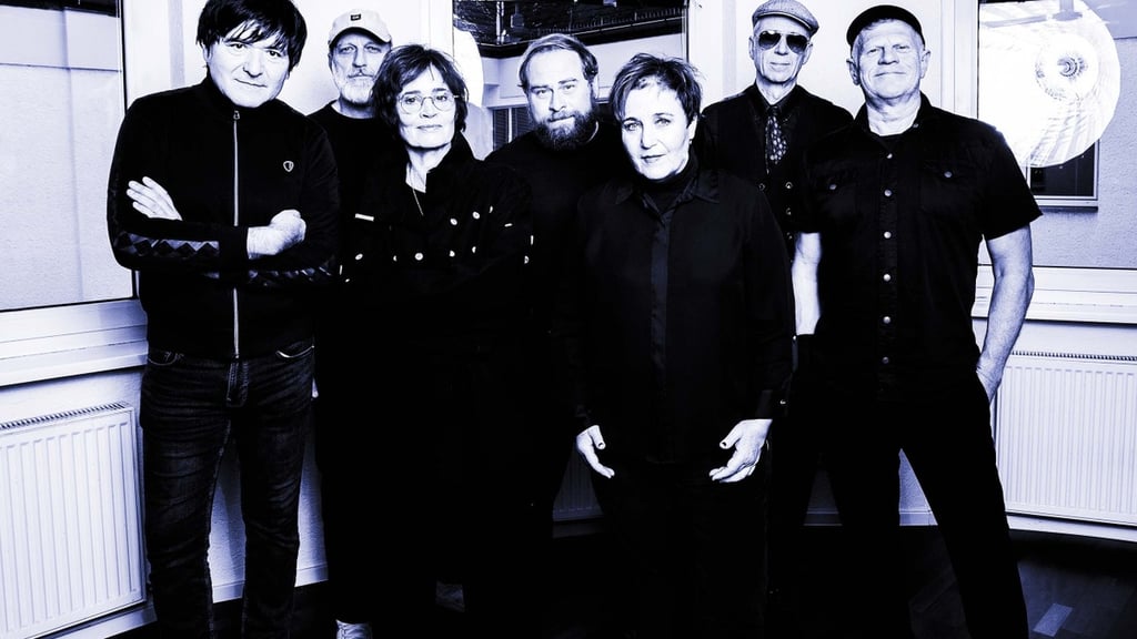 Group of eight adults in dark clothing posing in a recording studio, facing the camera.