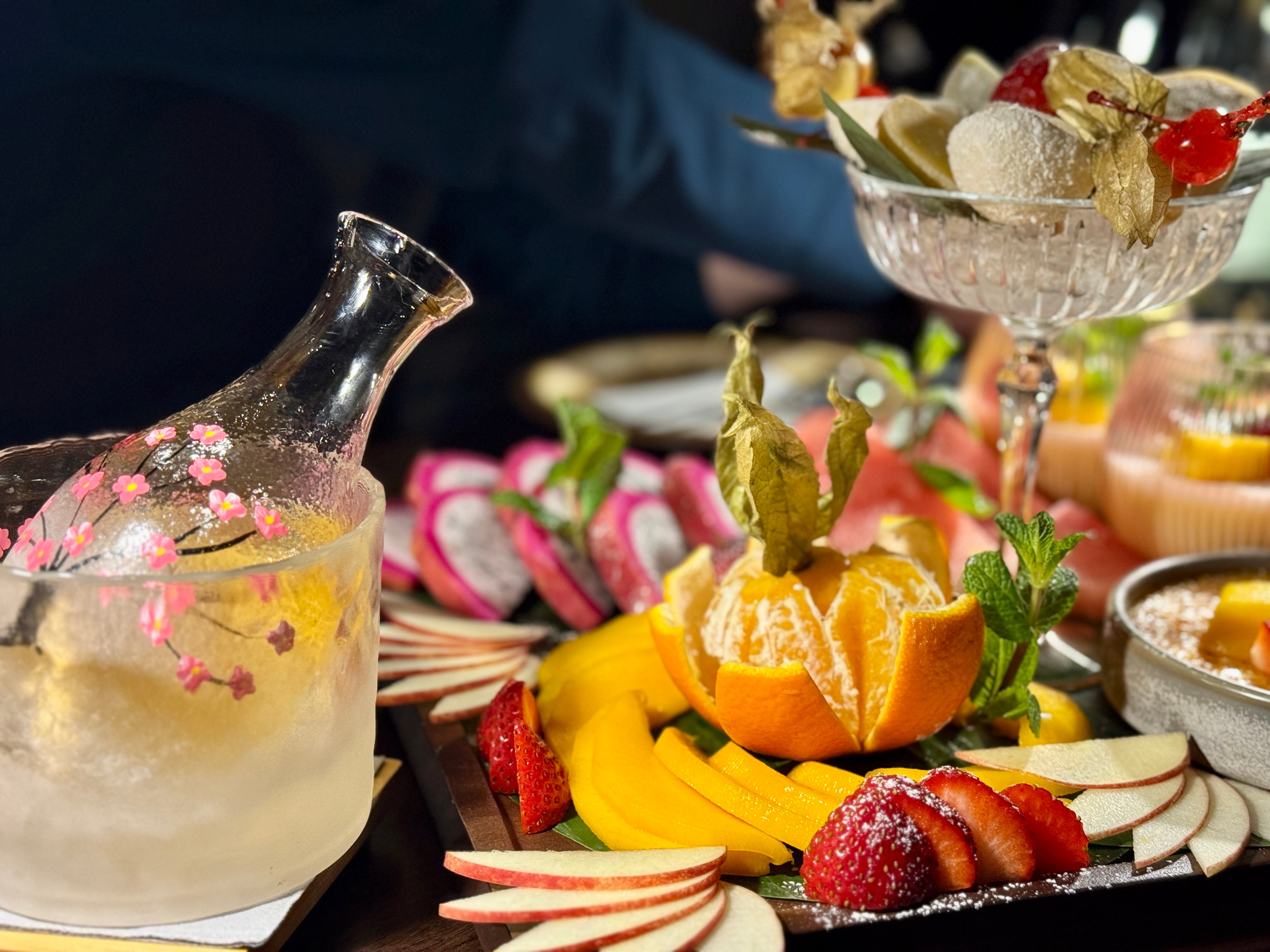 Dessertplatte mit Drachenfrucht, Orangenspalten, Mango, Erdbeeren und Apfel, dazu Mochi-Bällchen im Glas und eine Karaffe mit Eis