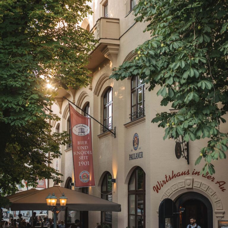 Beergarden exterior with arched windows, a Paulaner sign, and a red banner advertising beer and dumplings, sunlight through trees over outdoor seating under a beige umbrella.