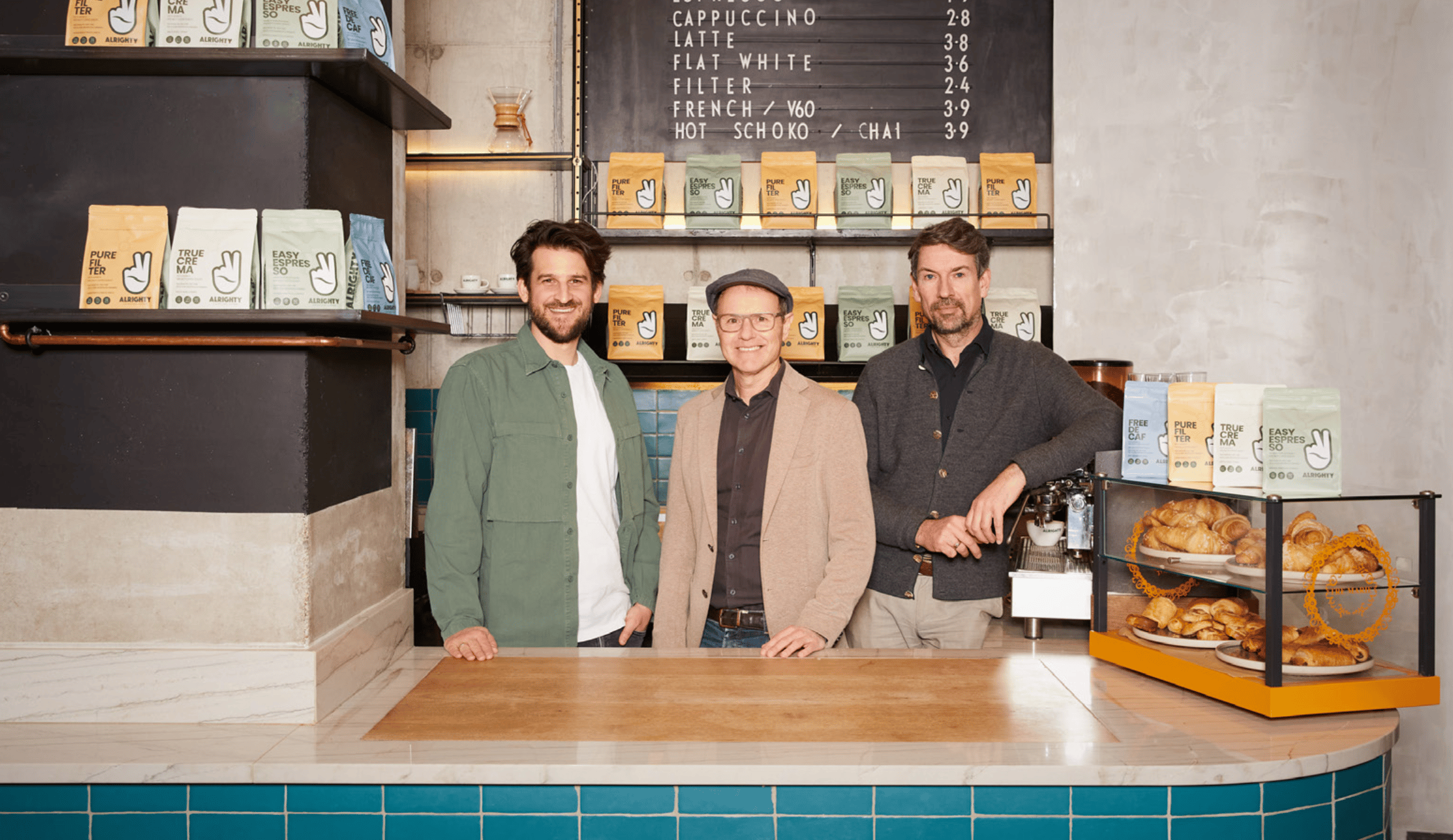 Die drei Gründer von Alrighty hinter der Theke des Coffee Shops mit Kaffeepackungen im Regal und Croissants in der Vitrine