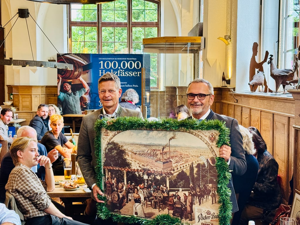Two men in suits pose with a large framed historical panorama, decorated with garlands, in a crowded Bavarian-style beer hall.