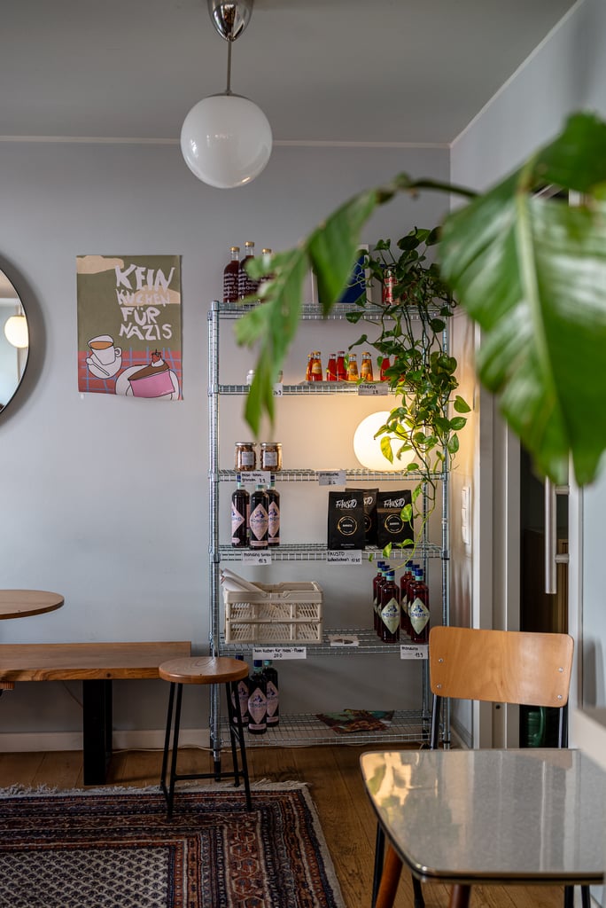 Interior of a cafe with a metal shelving unit stocked with bottles and jars, green plants draping over the shelves, gray walls, and a round poster reading 'Kein Küchen Für Nazis' on the left