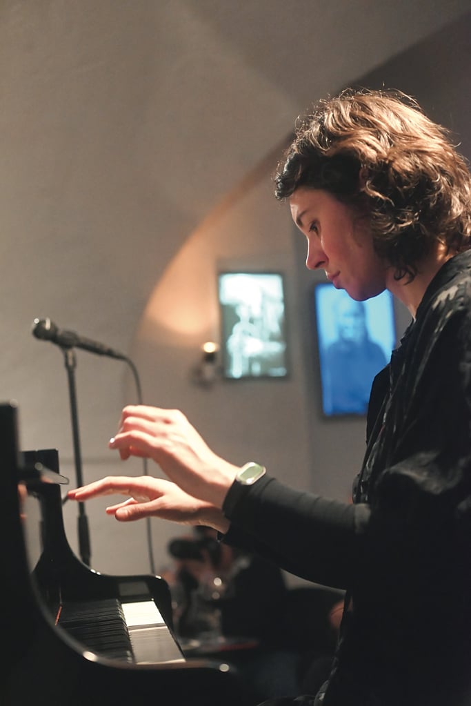 Musician in profile playing piano, hands on keys in a dim room with a microphone stand nearby.