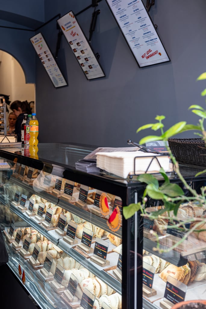 Bakery display counter with glass case filled with assorted pastries and labeled prices on small signs, blue wall behind.