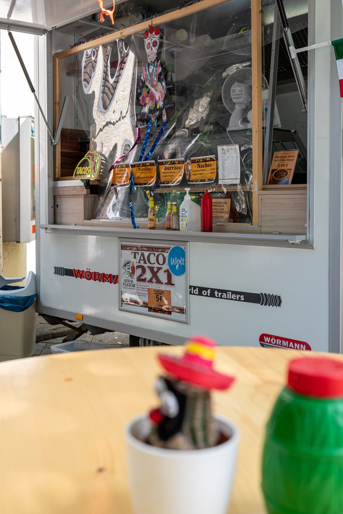 Food truck window with Mexican decorations and a taco/burrito/nachos menu; foreground shows a small potted cactus on a table