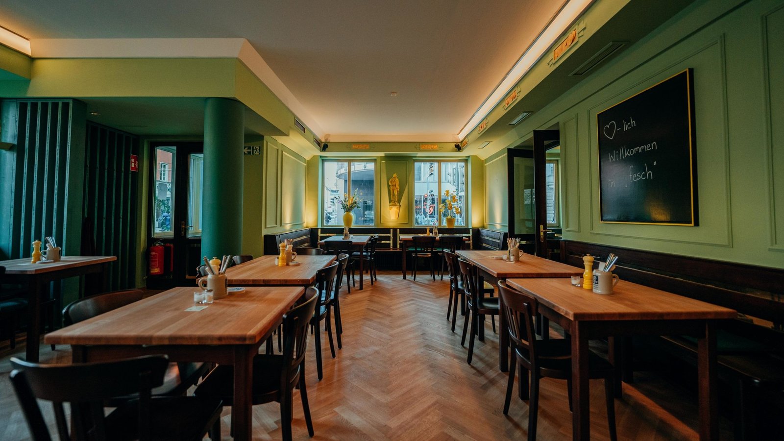 Hauptraum des Wirtshaus fesch mit grünen Wänden, Holztischen, Fischgrätparkett und Tafel