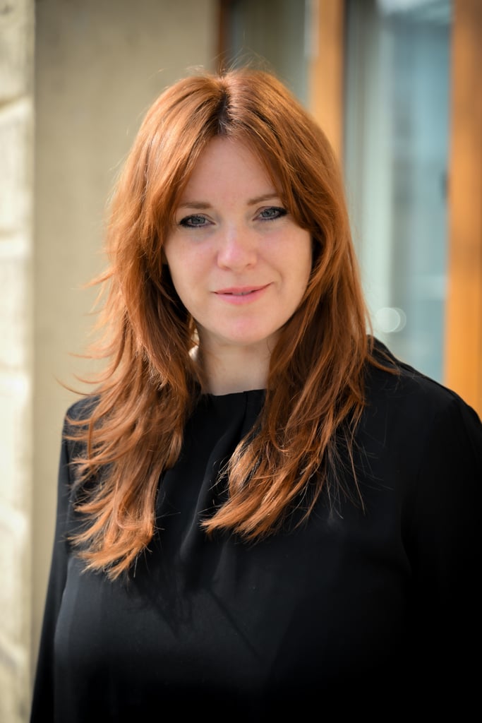 Close-up portrait of a woman with long red hair wearing a black top, smiling slightly outdoors against a blurred urban background.