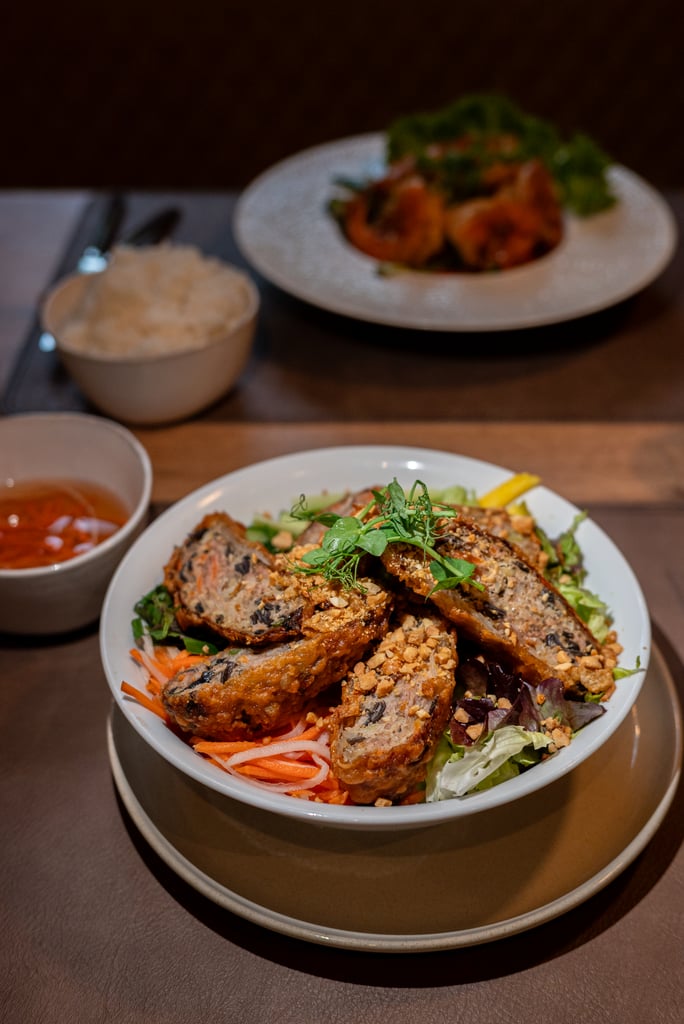 Crispy fried chicken pieces atop a bed of greens and shredded vegetables, garnished with chopped peanuts and fresh herbs on a white plate. Behind it, a bowl of white rice and a dipping sauce sit on the table.
