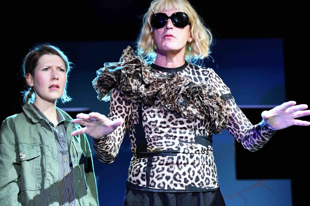 Two performers on a stage; the person in leopard print with large sunglasses and ruffled collar stretches their arms while the second performer in a green jacket looks on with a surprised expression.