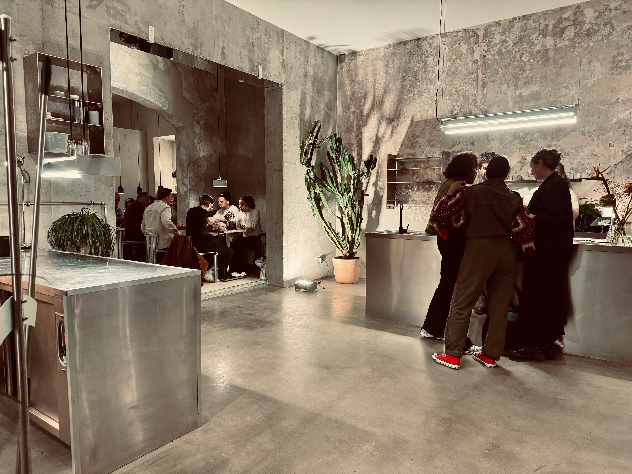 Gastraum des Nari Pizza Atelier mit grauen Steinwänden, Edelstahlflächen, Pflanzen und Gästen
