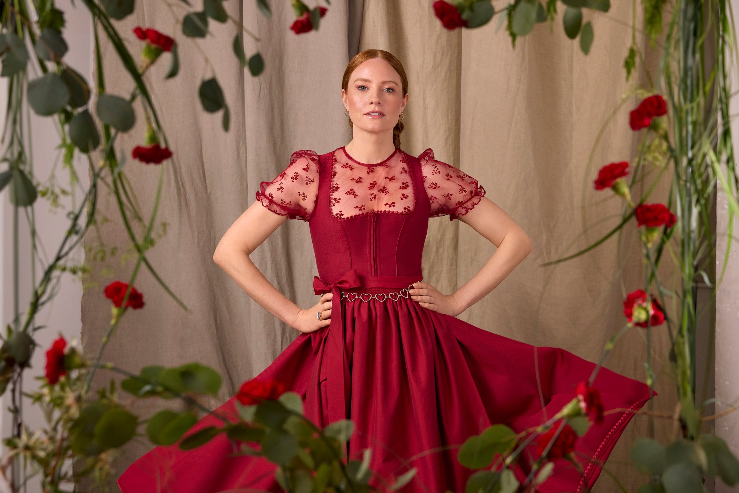 Portrait of a woman in a burgundy lace bodice dress posing with hands on hips among hanging greenery and red flowers.