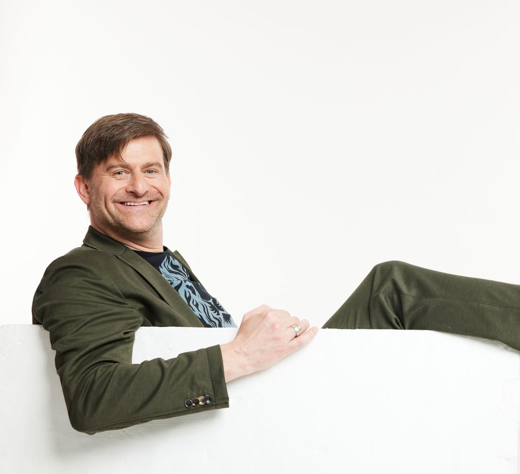 Smiling man in an olive blazer leaning on a white railing against a plain light background.