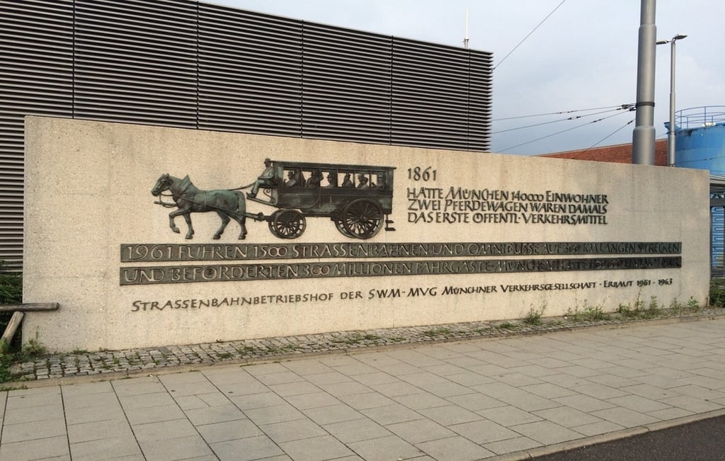 Stone monument with a bronze relief of a horse-drawn coach and passengers, accompanied by German inscriptions about early public transport history nearby.