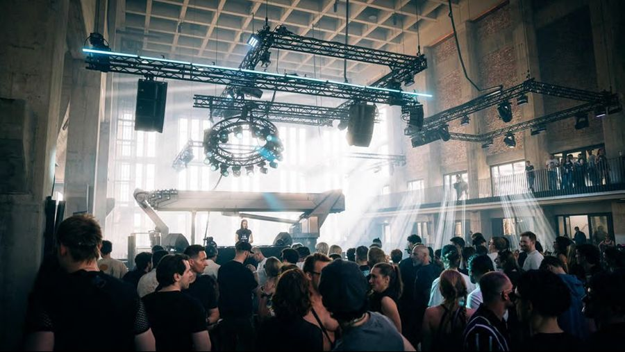 Crowd of people dancing in a dim club with a raised DJ booth and a circular lighting rig overhead.