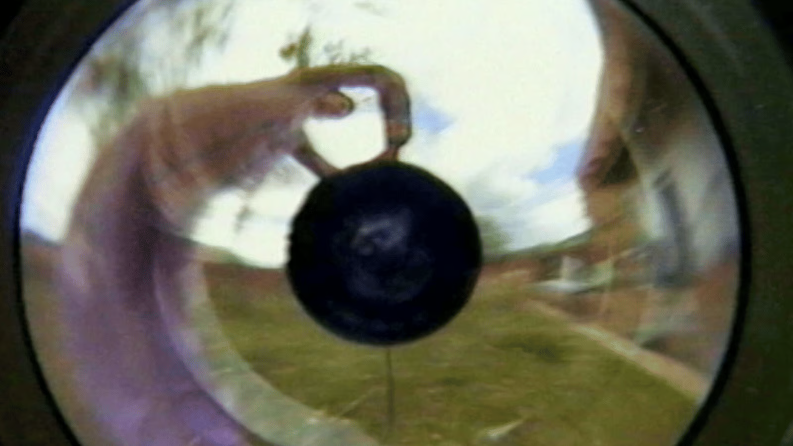 Close-up through a circular lens showing a hand reaching toward the camera outdoors in a park scene.