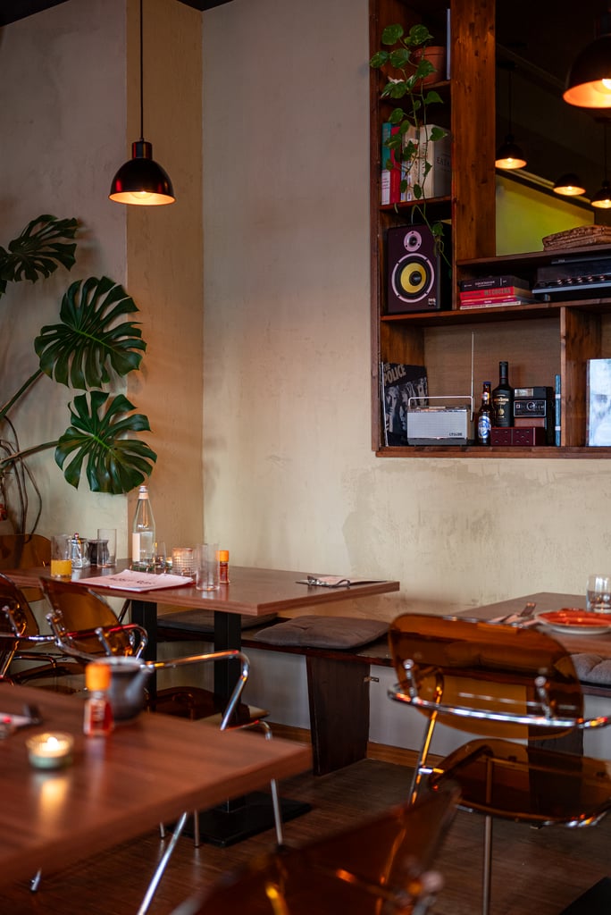Cozy cafe interior with wooden shelves, potted plants, and warm pendant lights over tables set for dining in the evening light.