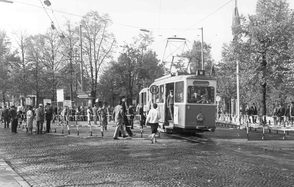 Vintage tram on a cobblestone street with pedestrians crossing and a crowd nearby, trees in the background. Readable signage and overhead wires indicate an urban street scene.