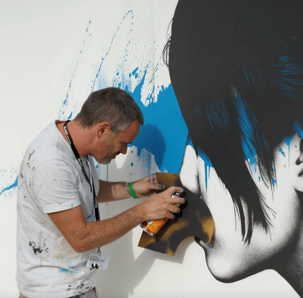 Man spray-painting a large mural on a white wall, holding an orange spray can in his hands