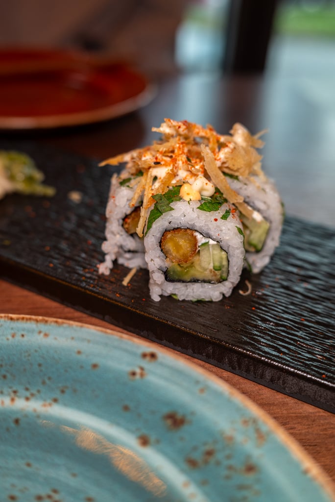 Maki sushi roll with avocado and cucumber, topped with crispy fried onions and chili flakes on a dark plate.