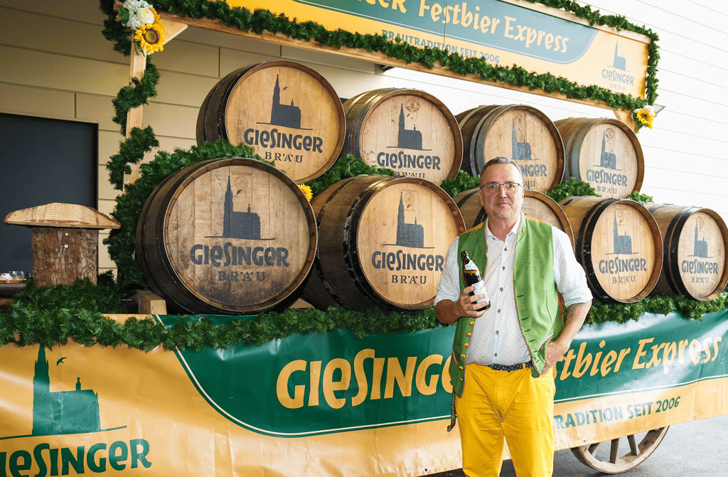 Man in a green vest and yellow pants holding a beer bottle in front of stacked wooden barrels labeled 'Giesinger Bräu' at a festival booth.