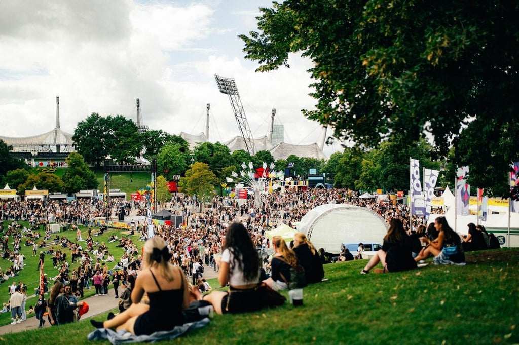 Large outdoor music festival in a park with people sitting on the grassy hill and a crowded festival area in the background, with stages and tents visible.