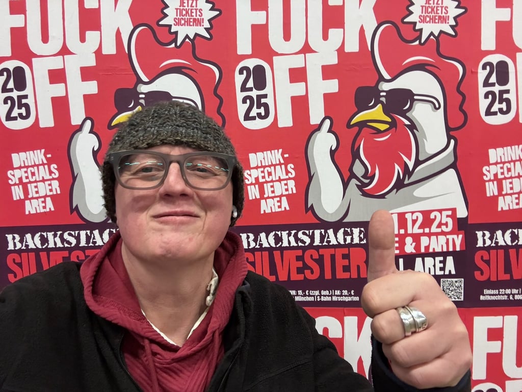 Person wearing glasses and a knit hat takes a close-up selfie in front of a bright red promotional poster with a cartoon rooster and bold white text, giving a thumbs-up gesture.