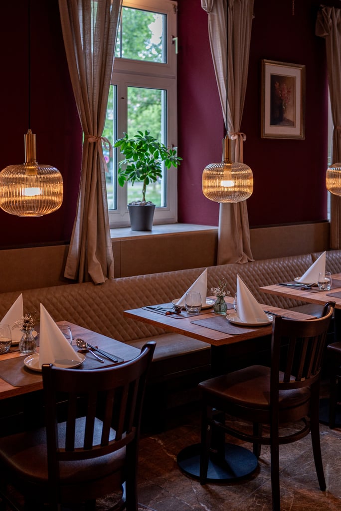 Cozy restaurant interior with wooden tables, folded white napkins, and brown chairs along a maroon wall; warm pendant lights.