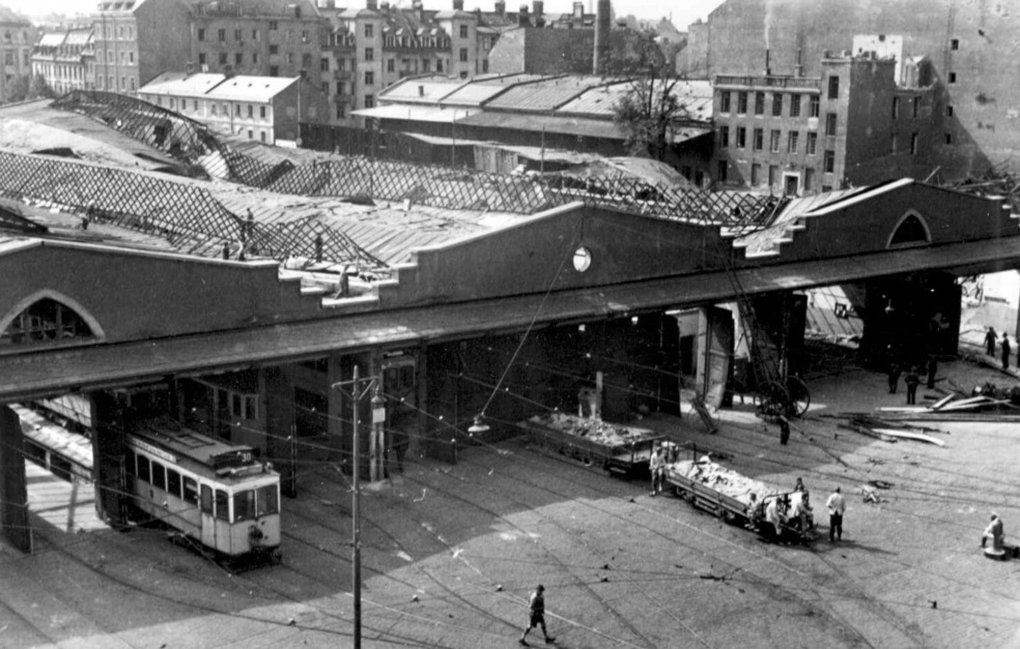 Black-and-white photo of a city square with a damaged elevated structure, a tram on tracks, and workers removing debris nearby.