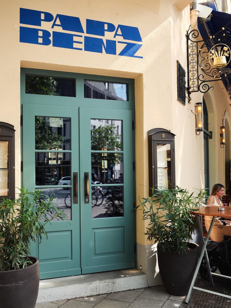 Cafe storefront with teal double doors, a bold blue 'PAPA BENZ' sign, potted plants, and a woman sitting at an outdoor table